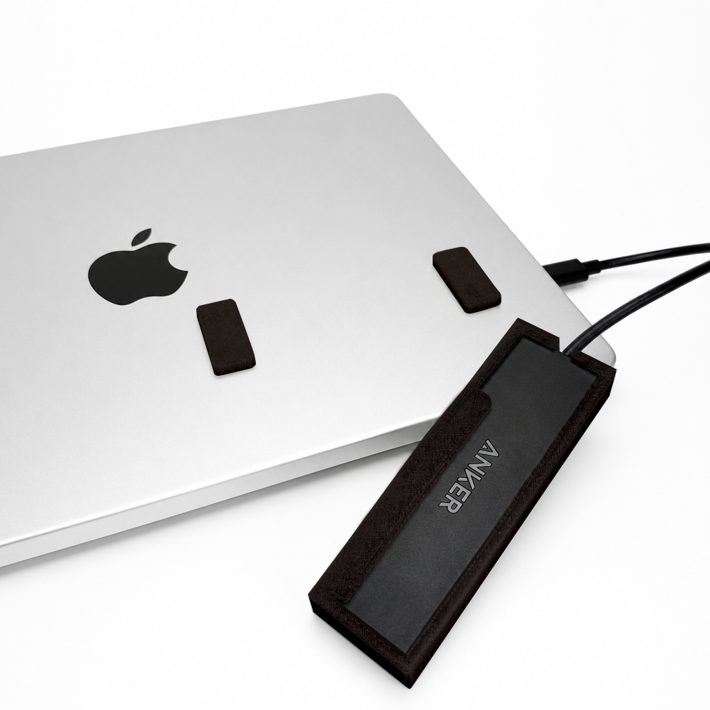 Front view of a black HubMount holding an Anker USB hub on a MacBook lid, with adhesive magnetic feet attached above the case.