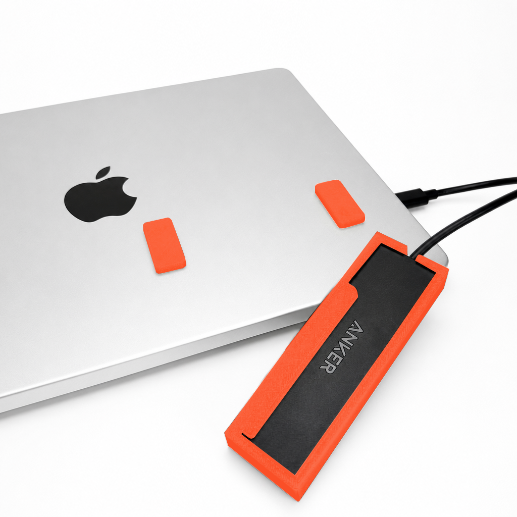 Front view of an orange-red HubMount holding an Anker USB hub on a MacBook lid, with adhesive magnetic feet attached above the case.